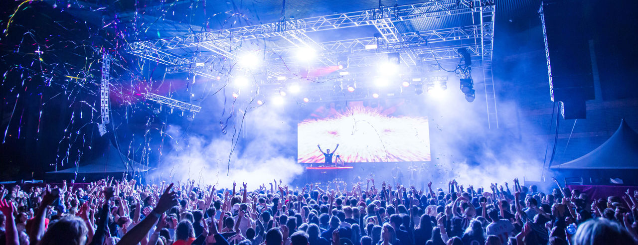 A man performs on a well-lit stage in front of a large crowd. Streamers and confetti fall from the ceiling. 